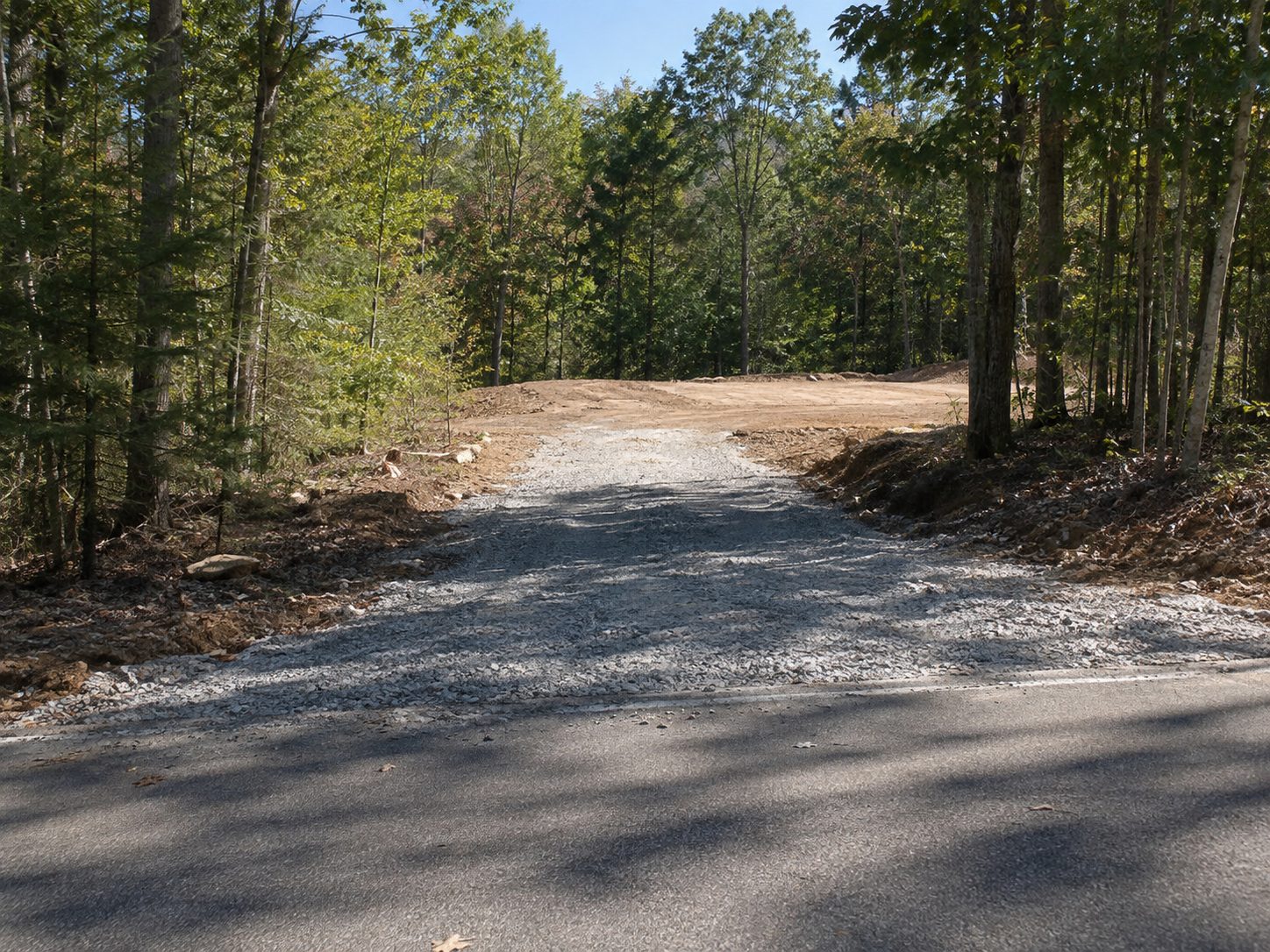 Gravel driveway cut heading back from a county road, with a fresh building pad or graded area at the end and woods on both sides
